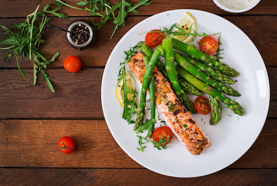 Top view of a baked salmon garnished with asparagus and tomatoes with herbs on a dark wooden table.