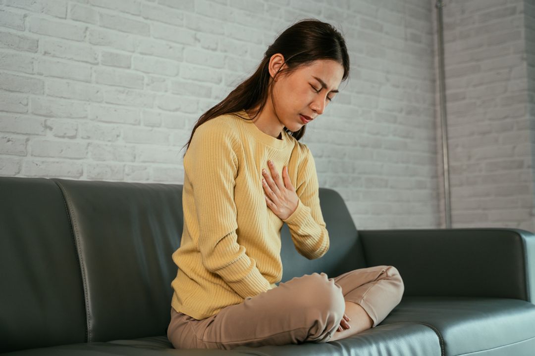 Woman sitting on a sofa crossed-legged with one hand on her chest in discomfort.