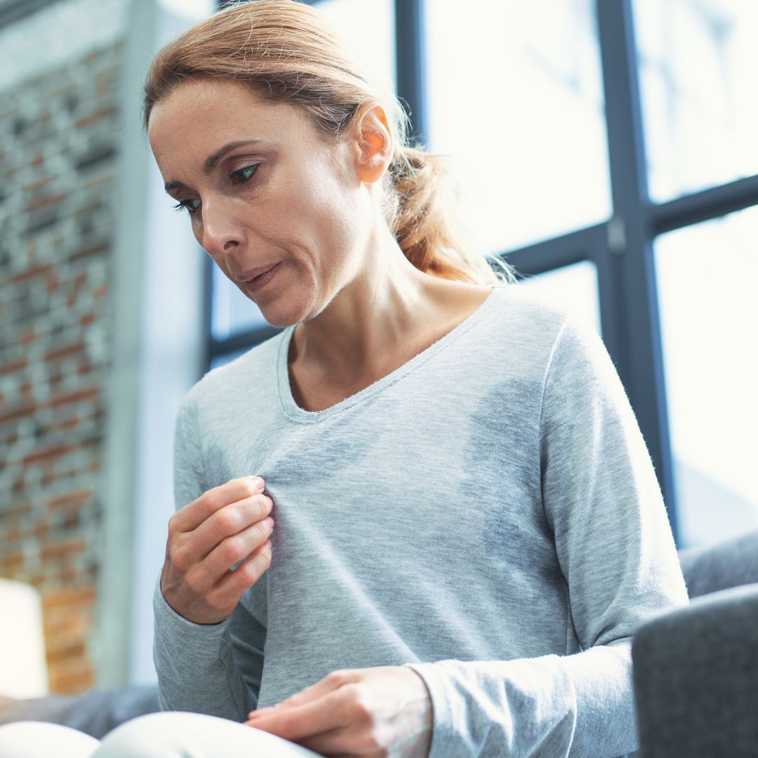 Woman touching her sweater and having a hot flash while sitting down on a sofa.