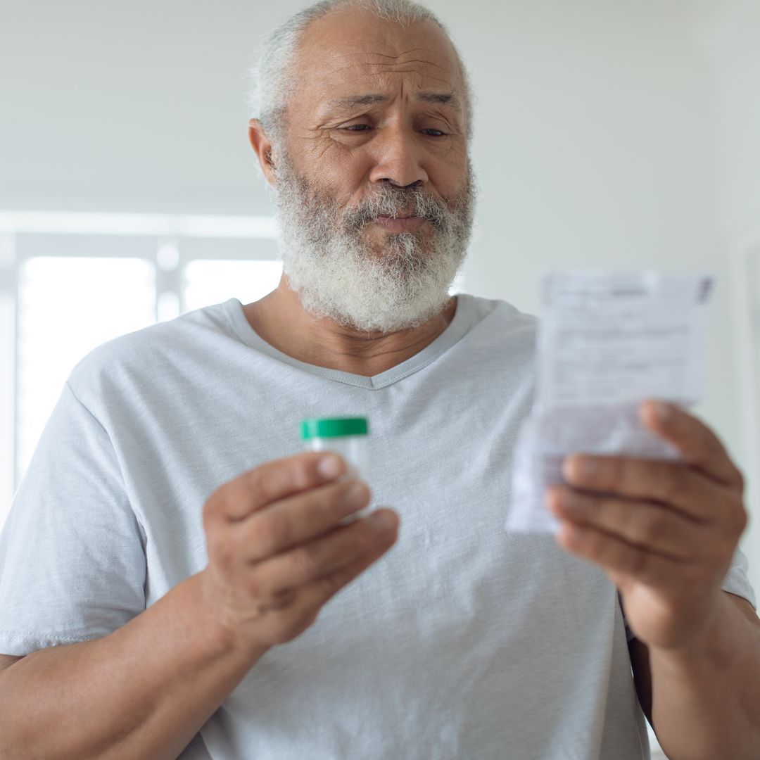 Man sitting on a bed holding a pill bottle while reading a prescription.