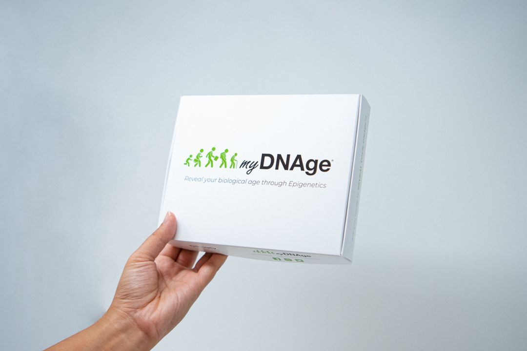 Hand holding a myDNAge test kit against a grey background.