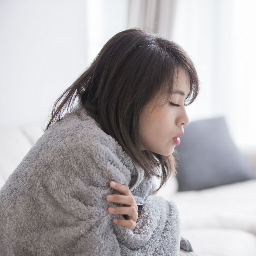A woman sitting on a sofa with her eyes closed wrapped in a warm grey blanket.