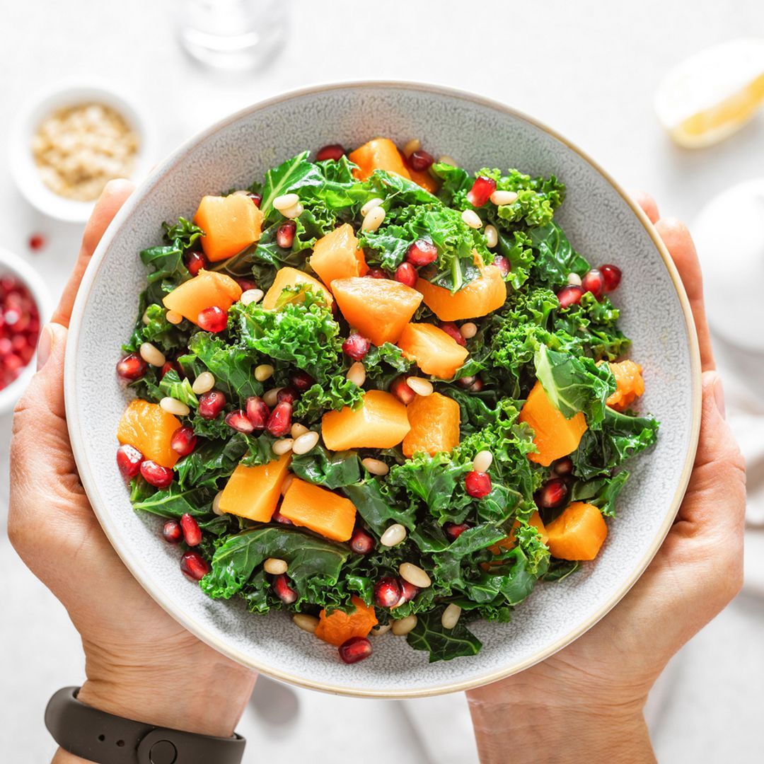 Hands holding a kale, pumpkin and pomegranate salad bowl over a white table cloth.