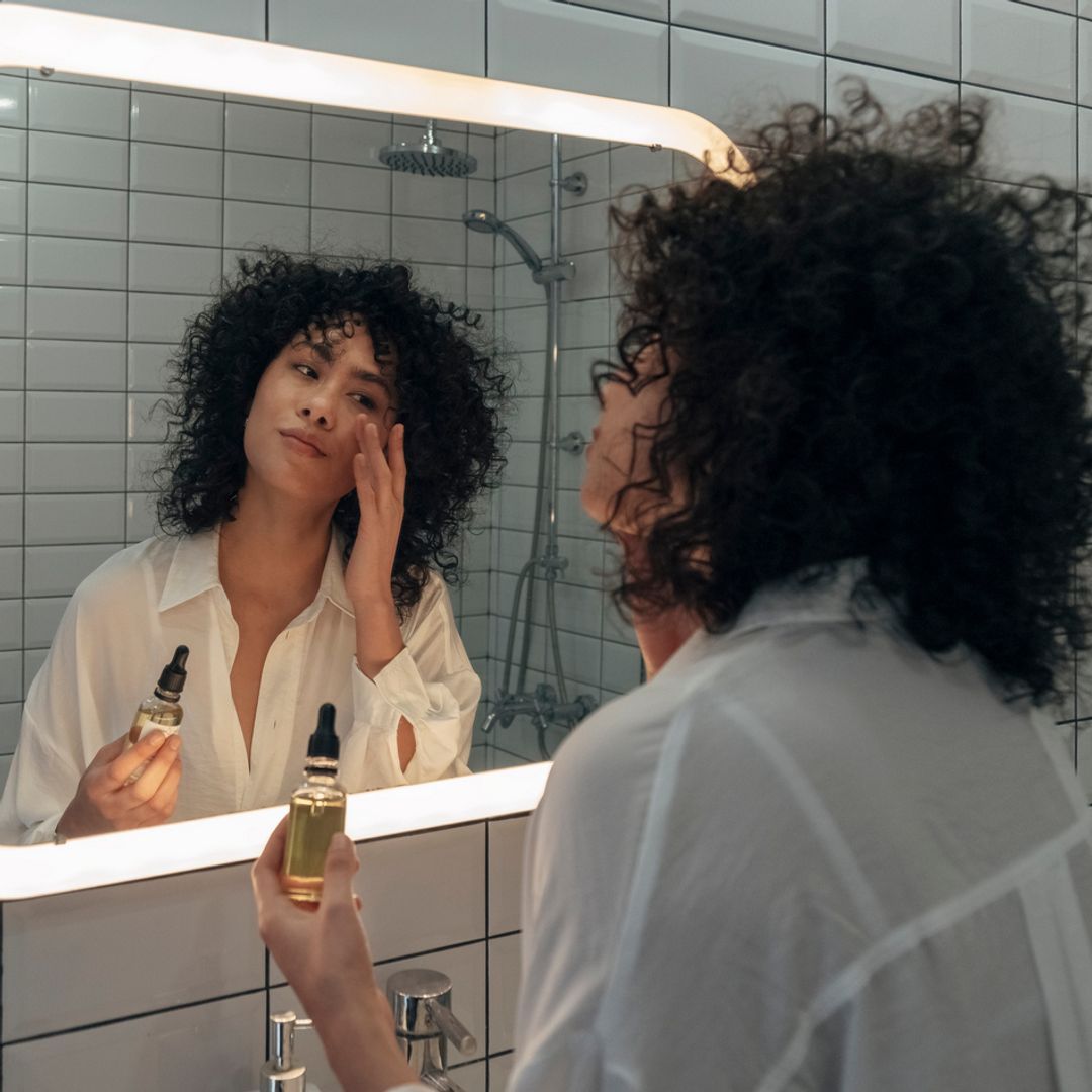 A woman standing in a bathroom looking into a mirror, applying a skincare product to her face.