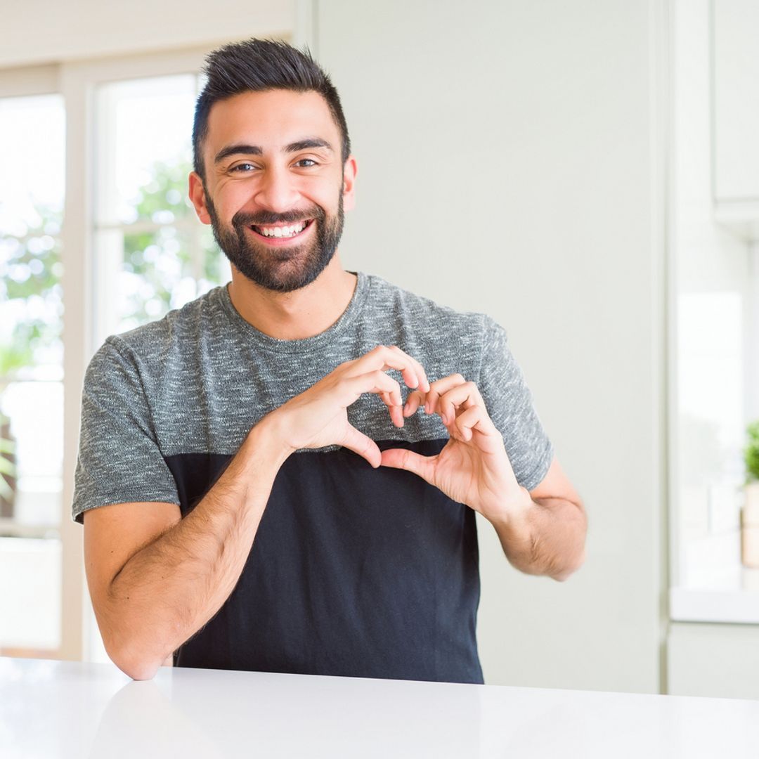  A man wearing a casual t-shirt at home smiling making a heart shape with his hands.