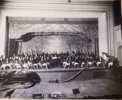 Black-and-white photograph of an orchestra posed on the auditorium stage, with a harp at the right and a piano in front