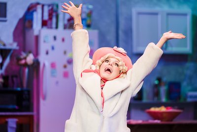 Tiger Drama performer in a pale coat and pink hat singing with arms raised