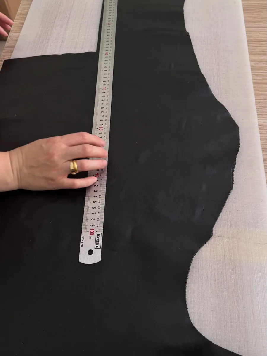 A hand holds a metal ruler over a piece of black material on the atelier worktable.