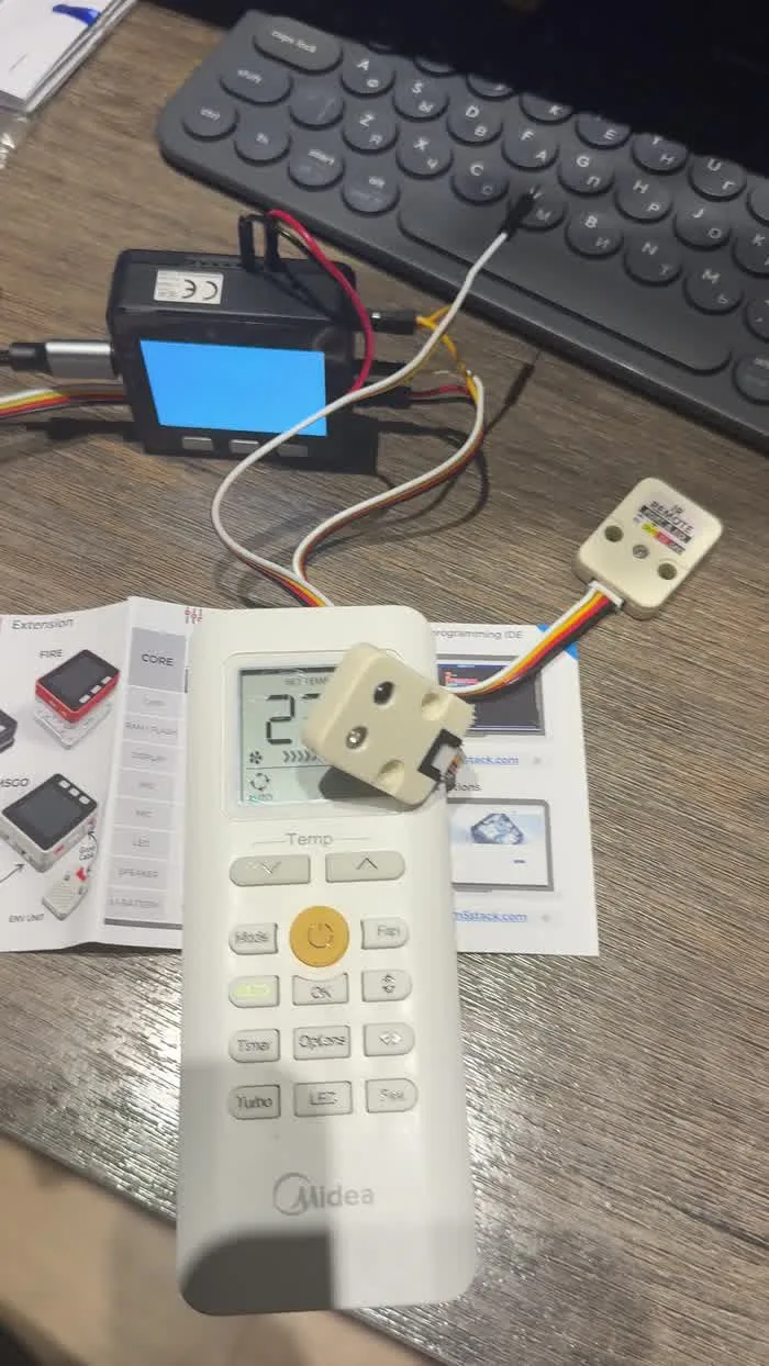 An infrared unit resting face down on an air conditioner remote on the desk, wired back to the controller, recording the codes each button press sends