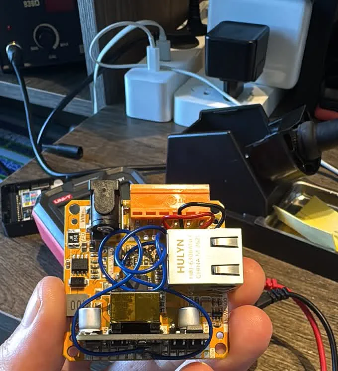 The PoE base held in one hand, four blue jumper wires soldered from its inner pads across to the orange screw terminal block, with a soldering station and multimeter on the bench behind
