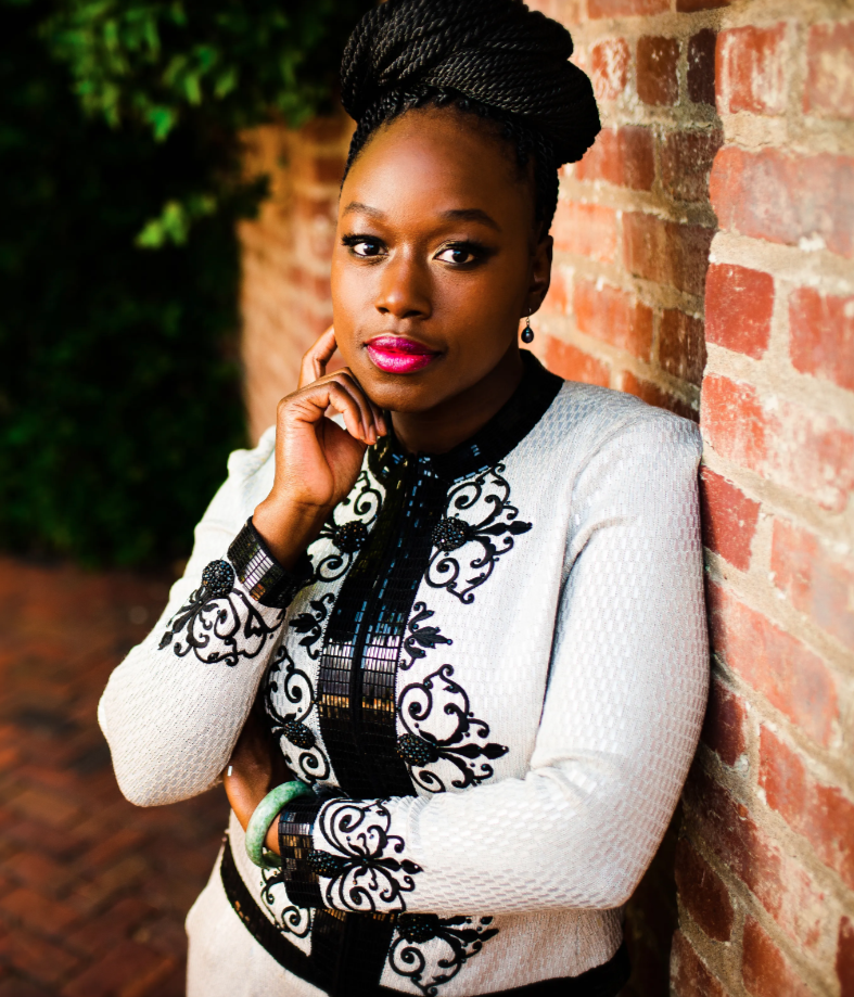 A photo of Angel, a Black woman with a bun in her head of long black braided hair. She is looking at the camera with a warm smile on her face and her eyes are sparkling.