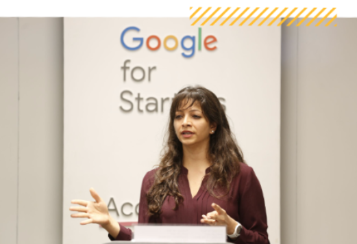 Wanita India berbicara di podium dengan logo Google for Startups di latar belakang.