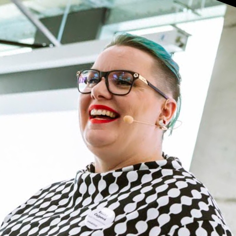 A photo of Eliška, a woman with short green hair and thick glasses. She is wearing a microphone in her head and is smiling while talking.