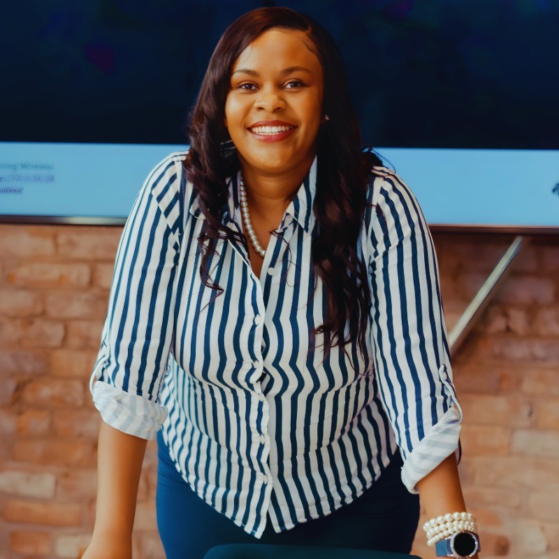 Madonna, a Black woman, stands in a power pose with a confident smile on her face. She is wearing a blue and white striped blouse.