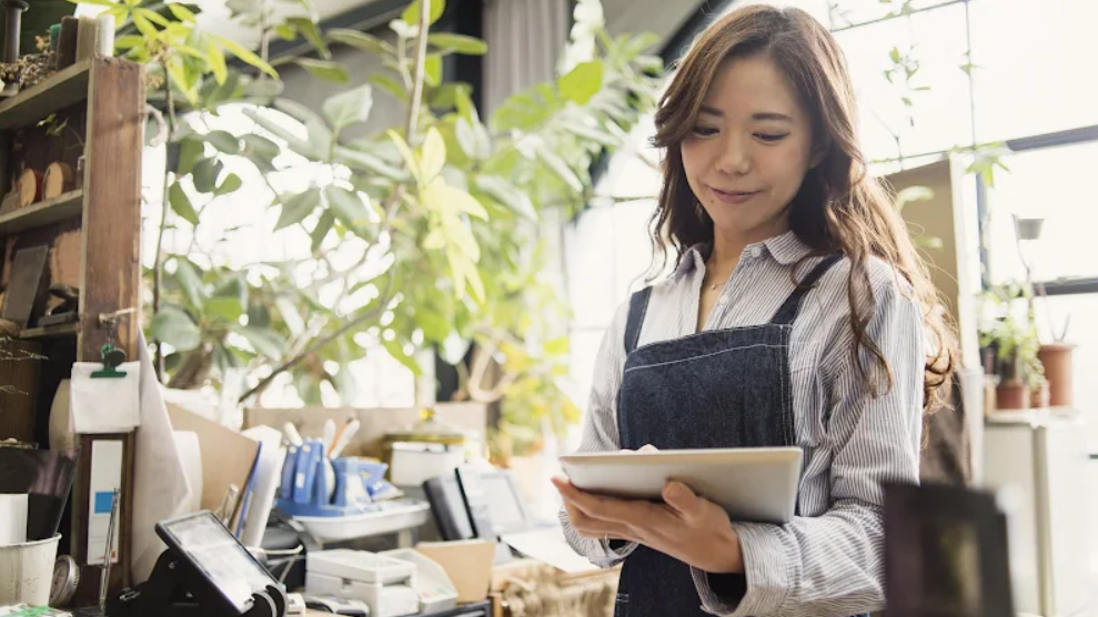 Una mujer examina una tablet de pie en una oficina llena de plantas con un escritorio y artículos de oficina.