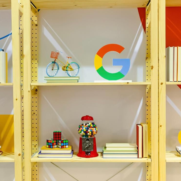 Полка с множеством разноцветных предметов. Миниатюрный велосипед ярких цветов выделяется среди классических головоломок, таких как кубики Рубика, игривый дозатор жевательной резинки и декоративная фигурка Google «G». Книги дополняют коллекцию, добавляя нотку знаний.