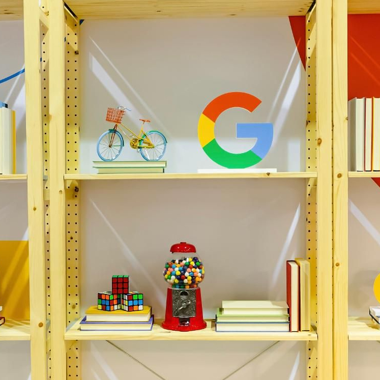 Rak yang menampilkan berbagai objek berwarna. Sepeda miniatur berwarna cerah tampil beda di samping teka-teki klasik seperti kubus Rubik, dispenser permen karet yang lucu, dan patung dekoratif Google &#39;G&#39;. Buku melengkapi koleksi, menambahkan sentuhan pengetahuan.