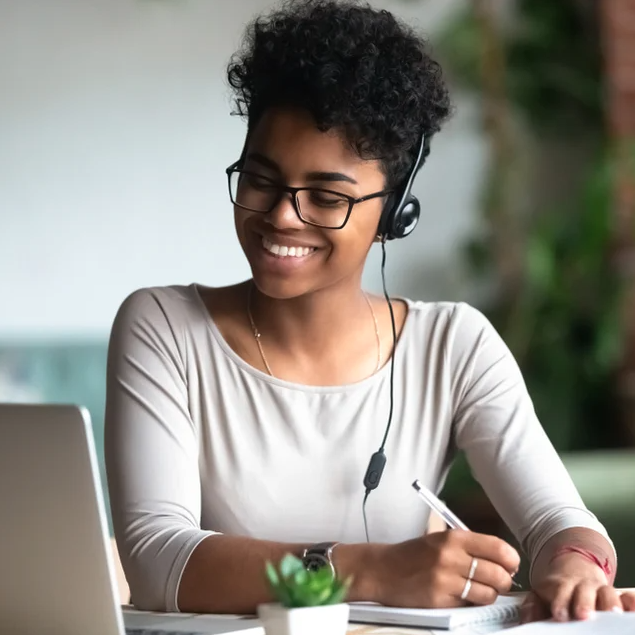A young Black woman with short hair and reading glasses is sitting at a table, looking at her laptop while writing in a notebook. She is smiling and wearing headphones. The photo captures her focused and creative energy as she works on her project.