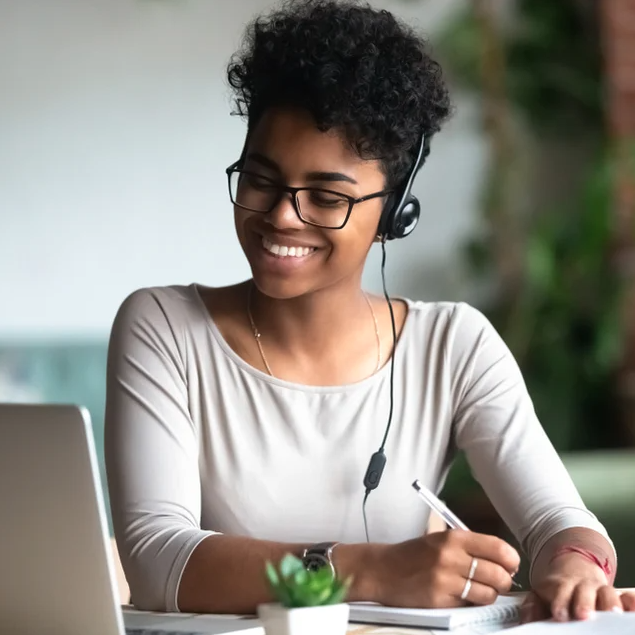 Una mujer negra joven con cabello corto y lentes para leer está sentada en una mesa, mirando su laptop mientras escribe en un cuaderno. Sonríe y usa auriculares. La foto captura su energía creativa y enfocada mientras trabaja en su proyecto.