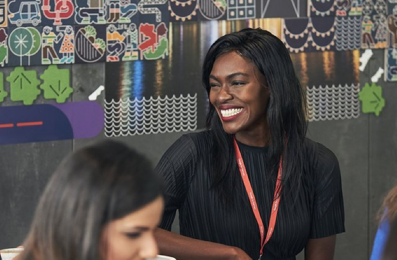 A photo of a Black woman wearing a black shirt smiling at someone off camera. She has long brown straight hair an exudes confidence.