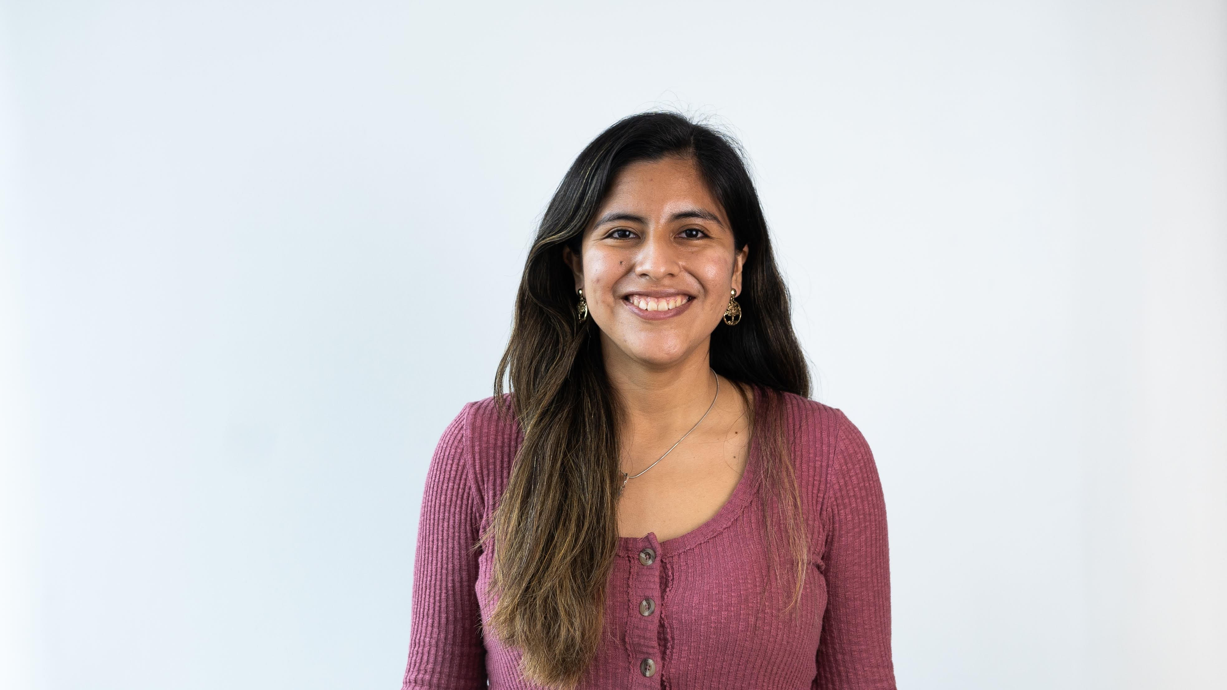 Victoria, a Peruvian woman standing in front of a white background. She's wearing a pink blouse and has her long light-brown hair lose while smiling at the camera.