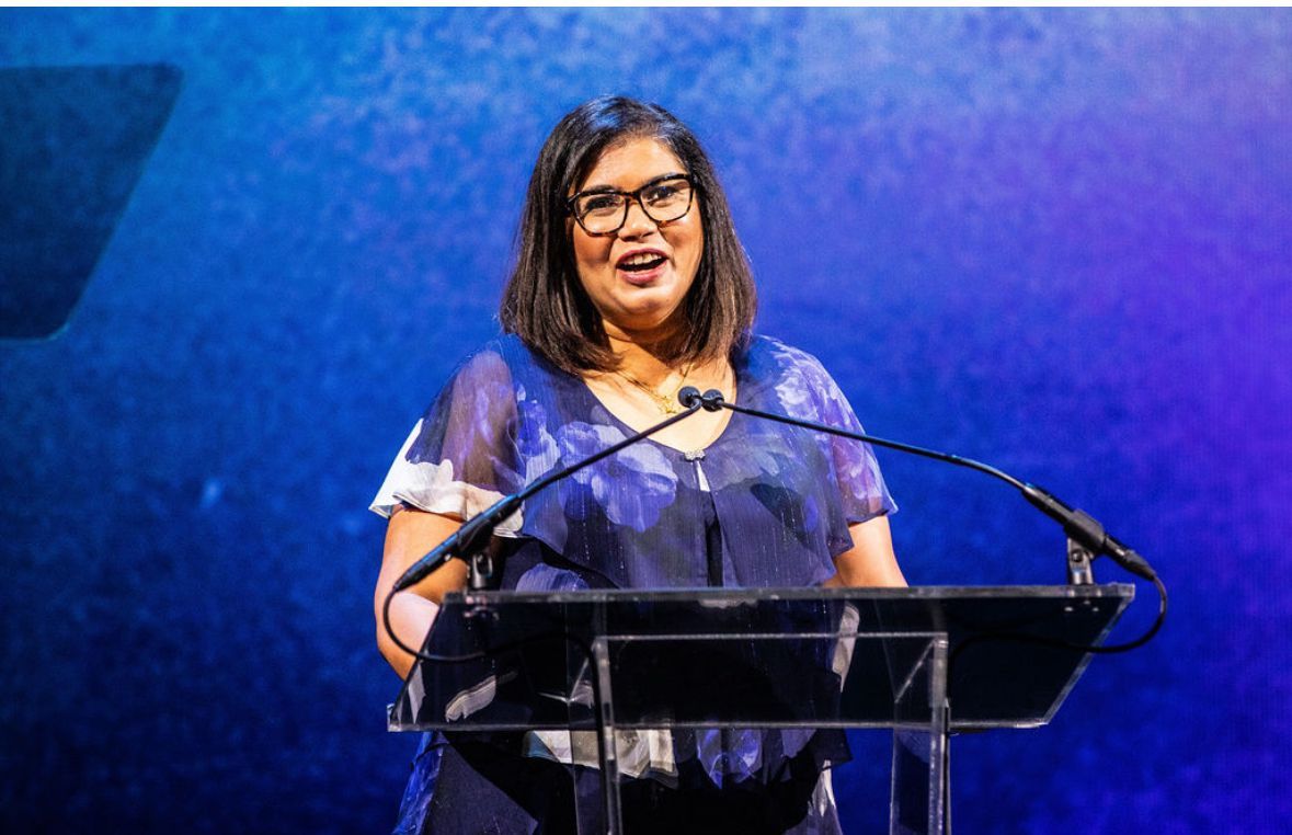 ElsaMarie speaks at a podium, wearing a blue dress and glasses.