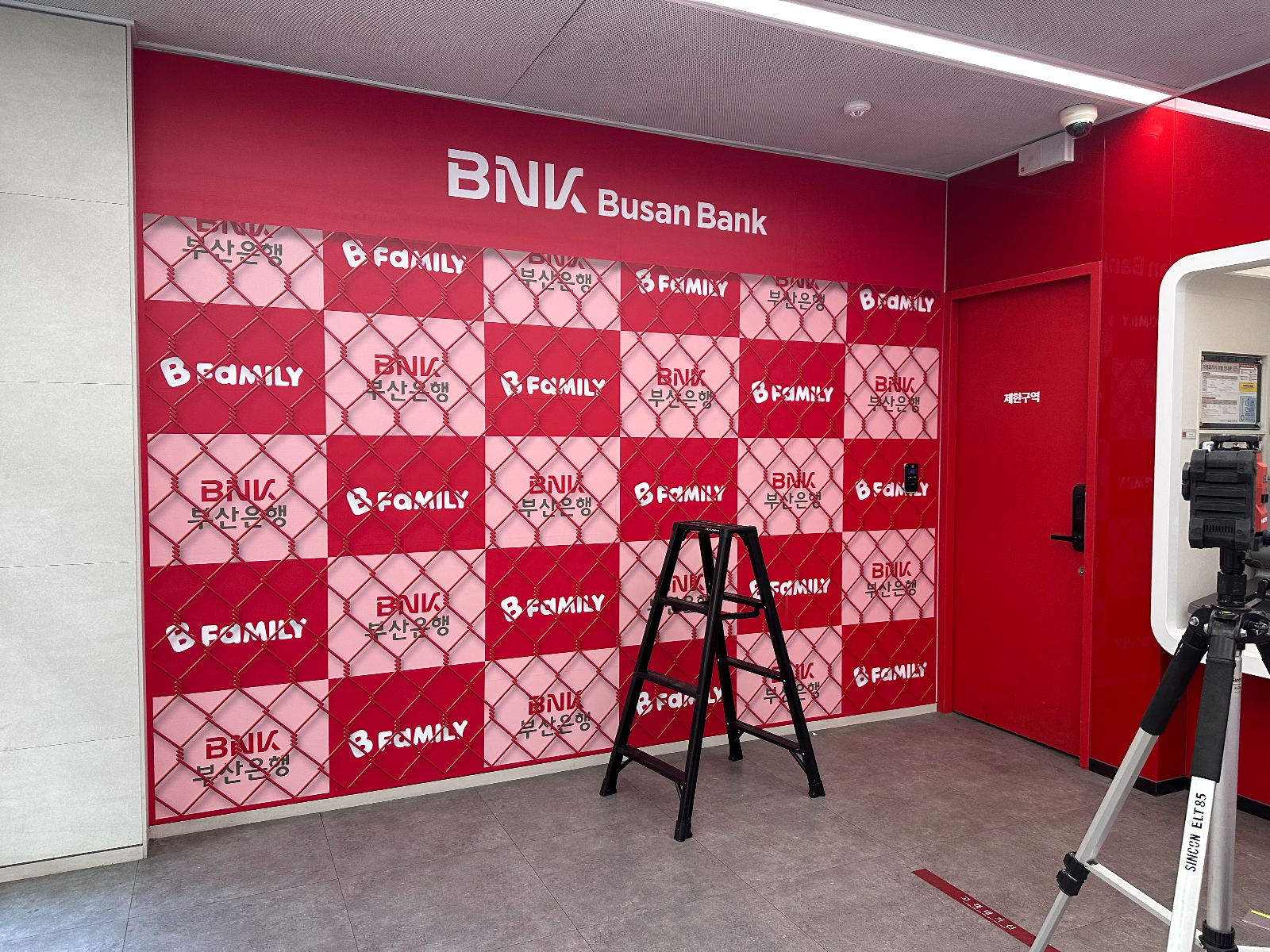 체크 패턴 랩핑이 끝난 벽면과 BNK Busan Bank 레터링, 앞에 놓인 사다리