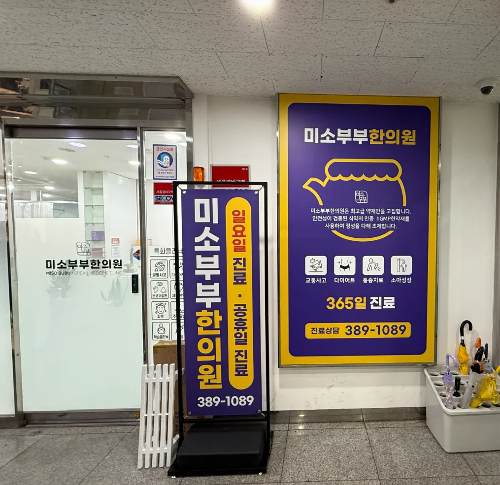 한의원 출입구 옆 벽면 게시물과 입간판, 출입문 유리에 상호 시트지가 붙어 있다