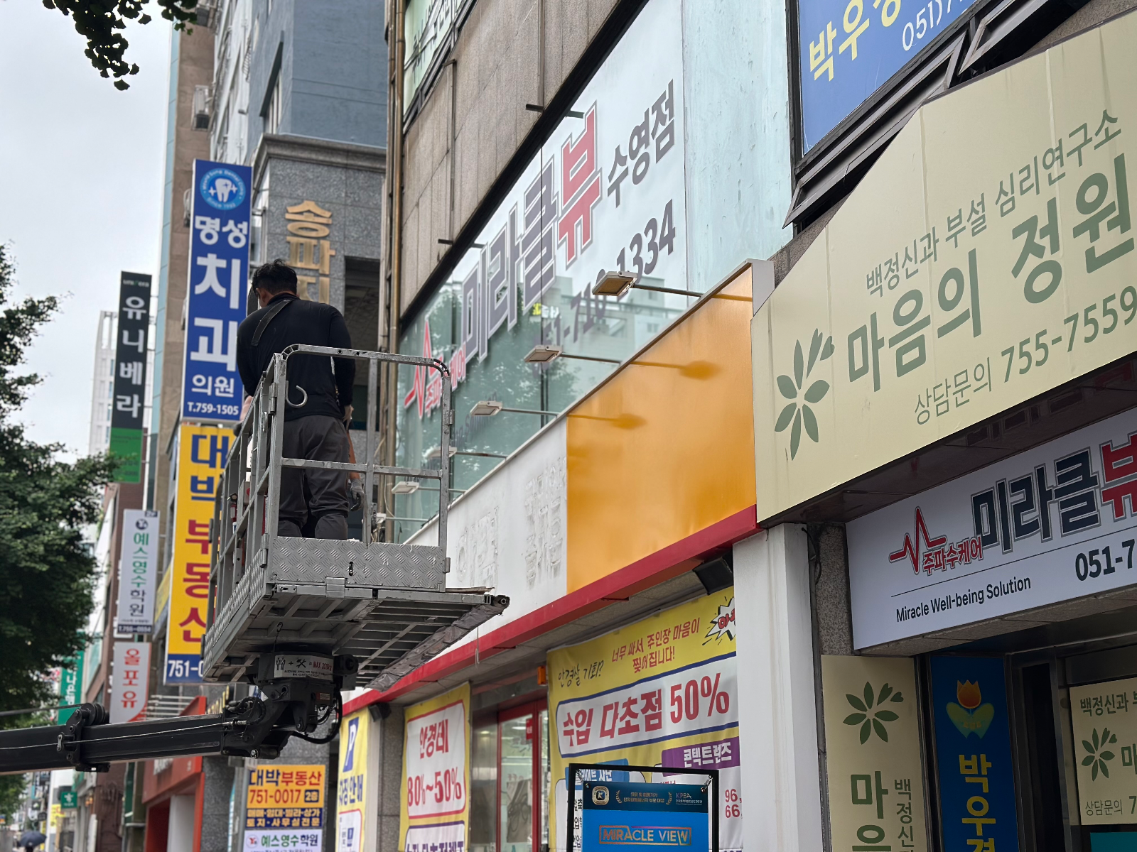 고소작업차에 올라선 작업자가 간판 프레임에 주황색 패널을 붙이고 있는 모습
