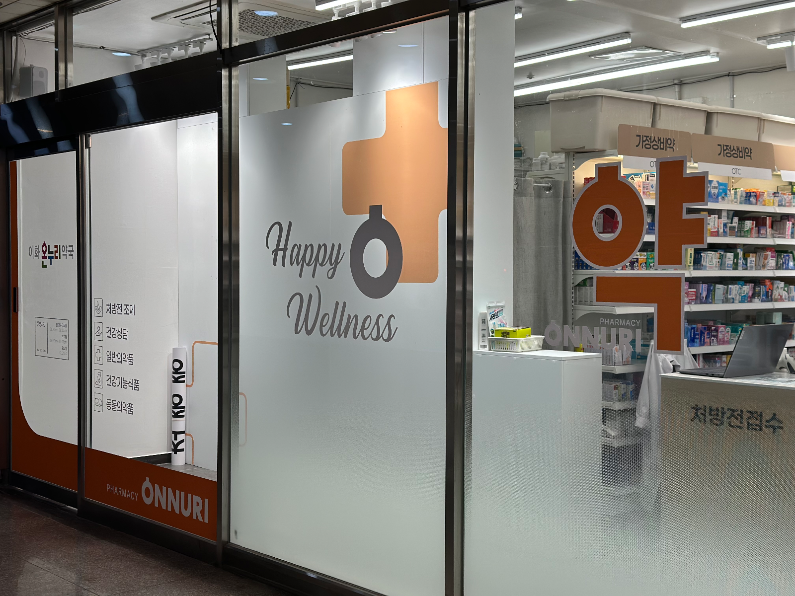 약국 내부 유리에 붙은 반투명 시트지. Happy Wellness 문구와 주황 십자 그래픽