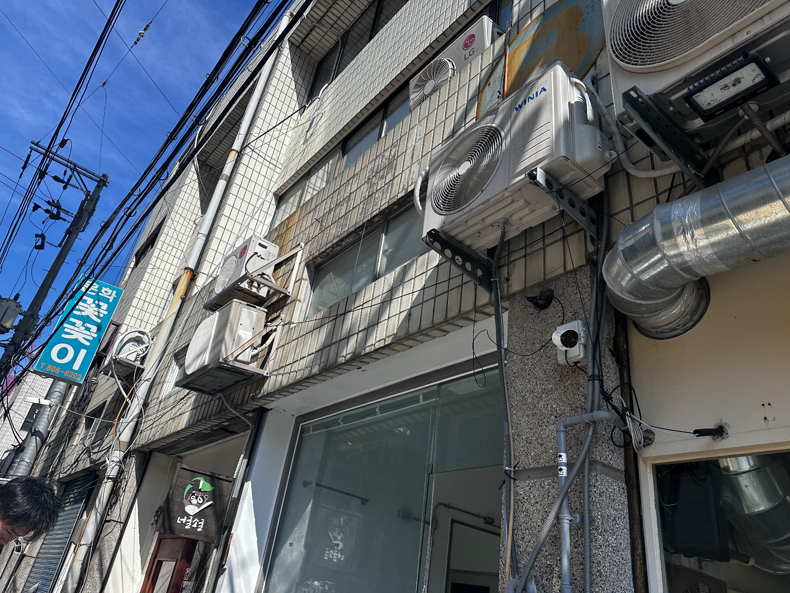 간판을 달기 전 건물 정면. 회색 타일 벽과 유리 파사드, 위층 에어컨 실외기가 보인다