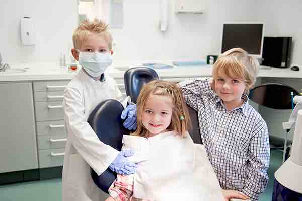 3 kids play acting at the dentist office, one is dressed like a dentist.