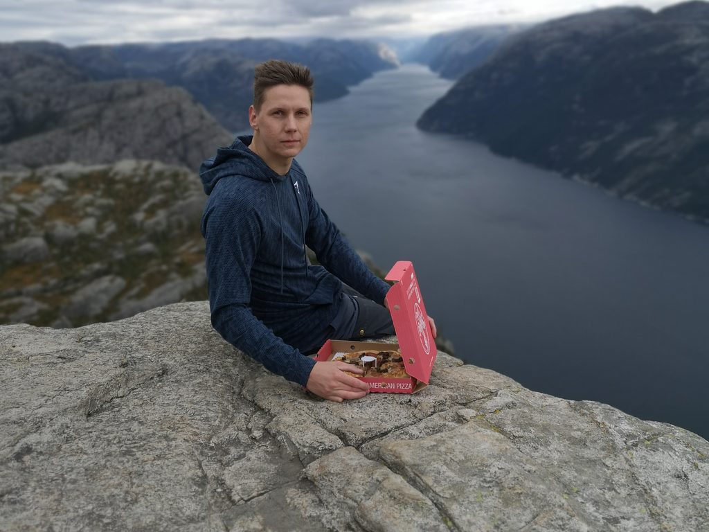 Helge sitter på kanten av Preikestolen med Lysefjorden bak seg og en åpen pizzaeske ved siden av seg.