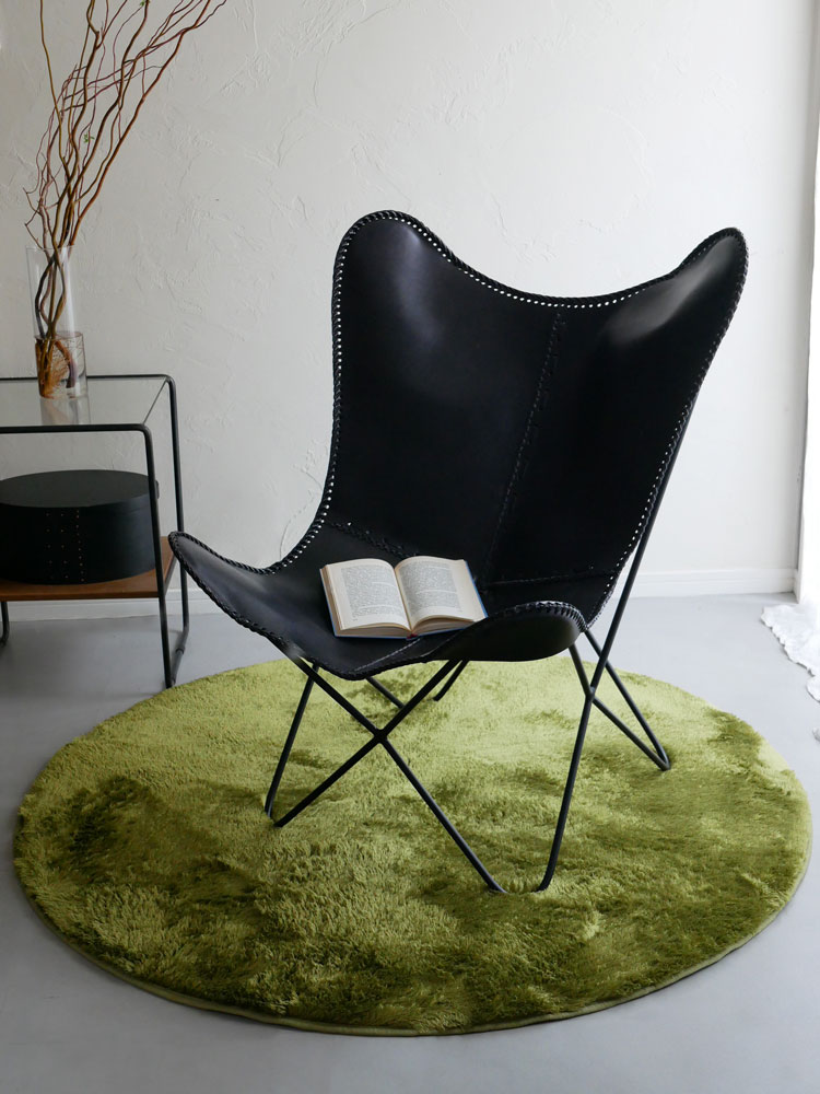 ポリエステル製130cm円形ラグが木製フローリングのリビングルームに敷かれ、クラブチェアやレザーソファとともに快適な空間を演出している様子