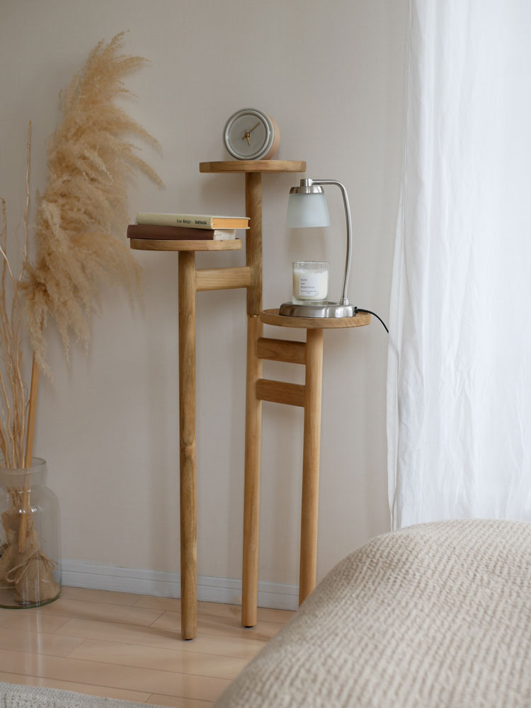 ナチュラルなチーク無垢材製サンフラワーラタンのステップトレイテーブル。高さ調節可能な3段トレイとシェルビングが配置され、ランプや花瓶、観葉植物が飾られたリビングルームのインテリアを演出しています。