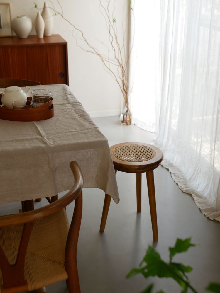 チーク材とラタンで作られたサンフラワーラタンのラタンスツールが、木製の床と花鉢が配置された室内インテリアに置かれています。