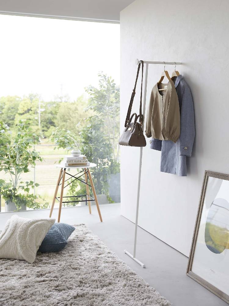 リビングルームの木製フローリングに設置されたスリムコートハンガー。スチール製でシリコーンのフックが特徴的。周囲にはランプや観葉植物、カーペットが配置されている
