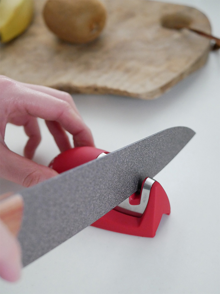 協和工業のスーパーストーンバリア包丁と専用シャープナーが切り板の上に配置され、果物や肉とともに調理風景を示しています。