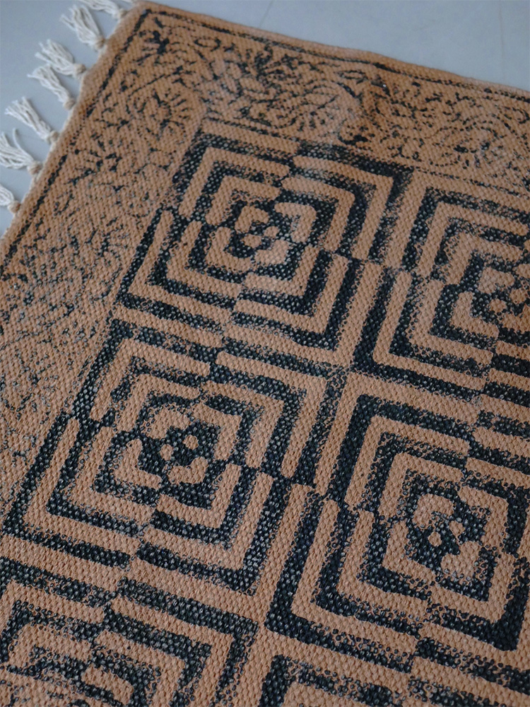 アデペシュ マニエール コットンラグ。茶色の対称的な模様が施されたウーブンファブリック製カーペットが床に敷かれています。