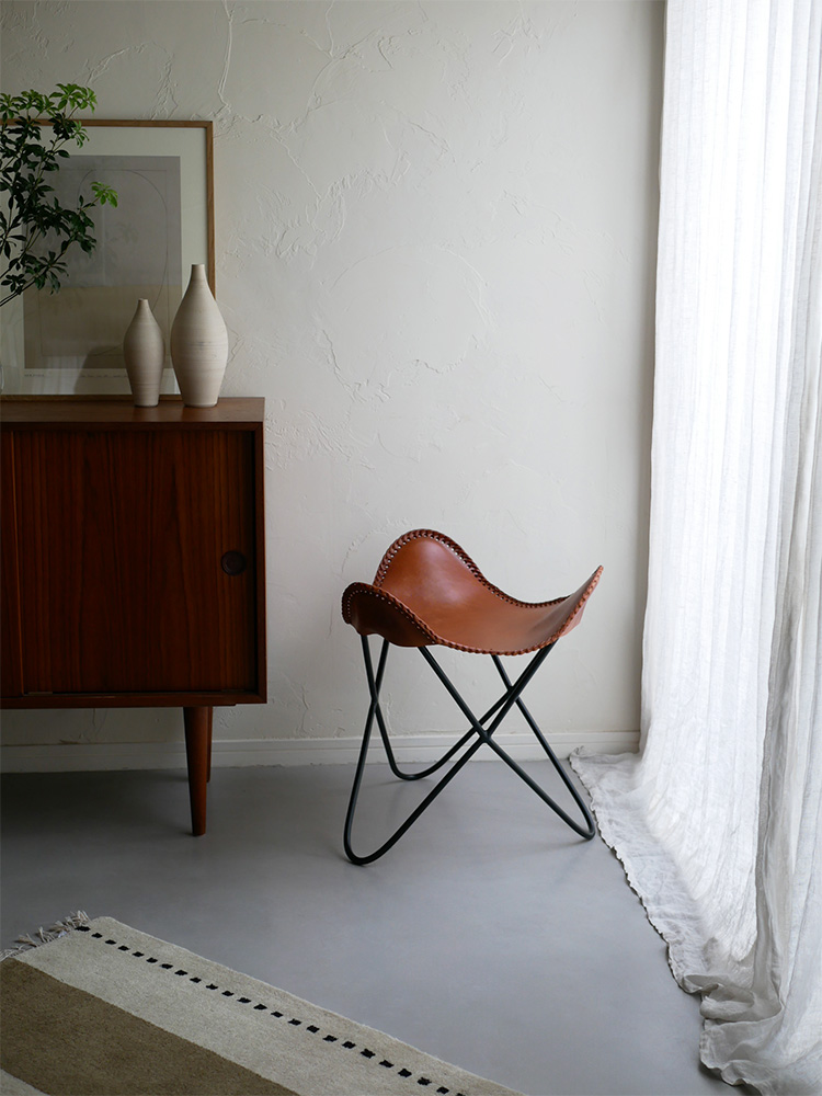 ブラウンの木製フローリングの上に配置されたWRAPSラップス アイアンレザースツール。牛革とアイアン素材で作られたモダンなデザインの椅子