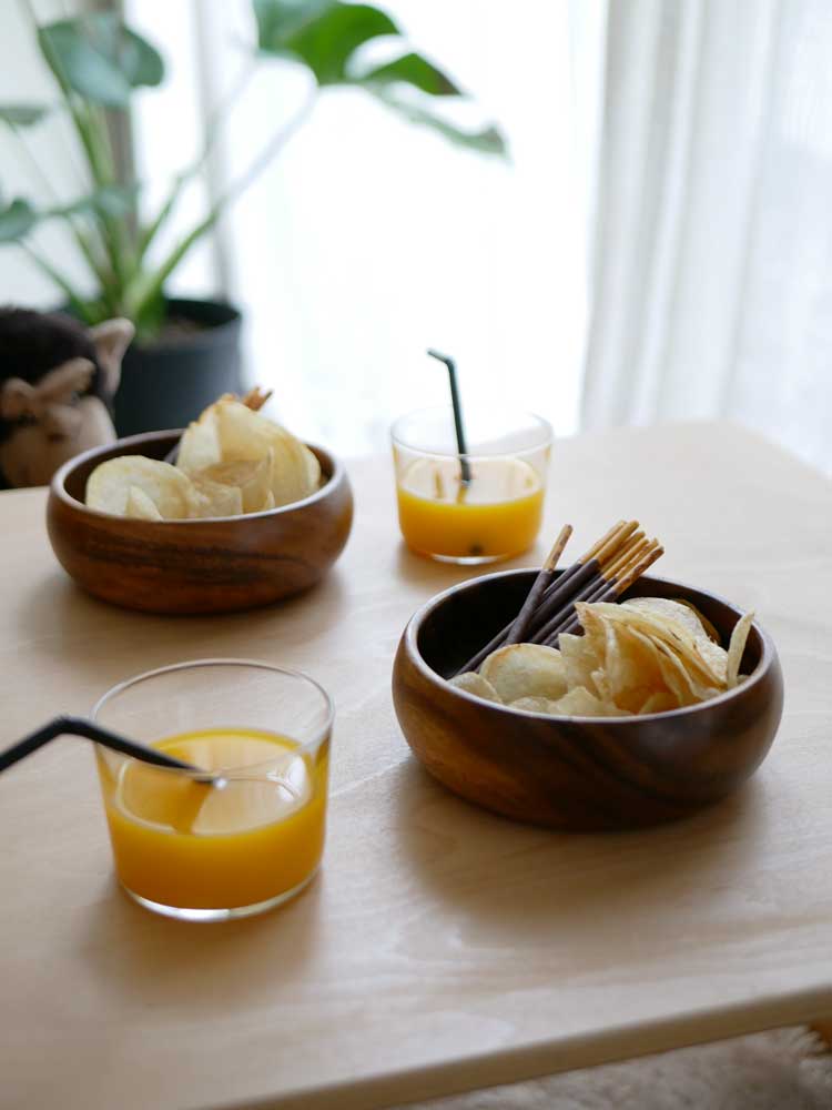 アカシア木製のエッグ型トレー、スクエアトレー、ラウンドボウルがオレンジジュースと共にテーブルに並べられている様子