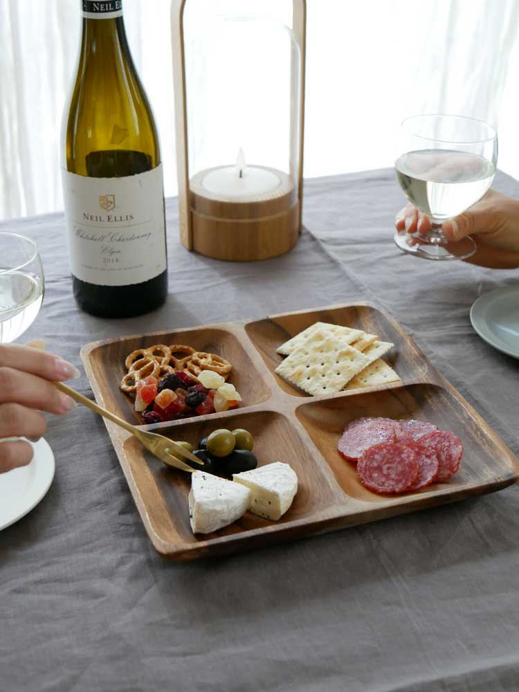 アカシア製の選べるやさしい食器セットがテーブルに並べられ、チーズやワインボトルを含む料理と共に配置されている様子