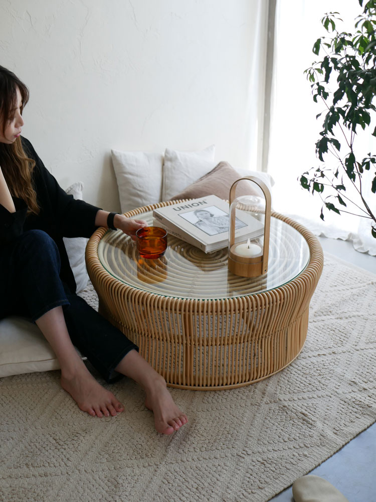 ラタンとガラスを使用したサンフラワーラタンのローテーブルが、ソファやクッションと共にリビングルームに配置され、ナチュラルで快適な空間を演出しています