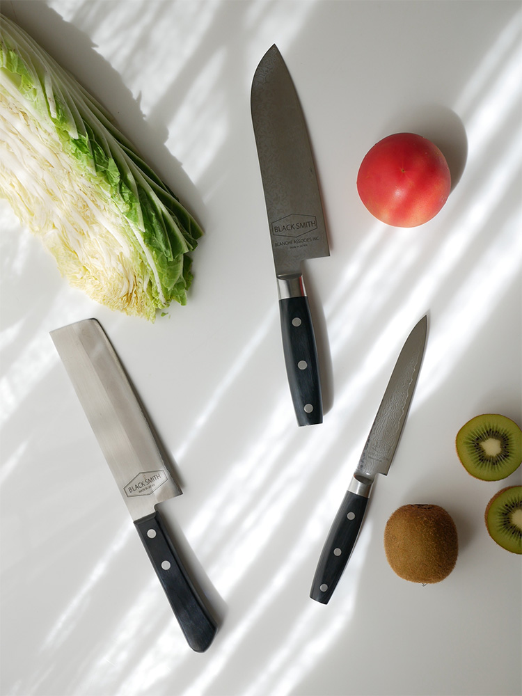 日本製TOOLSブラックスミスのダマスカス鋼キッチンナイフセットがカッティングボードと新鮮な野菜とともに配置されている様子