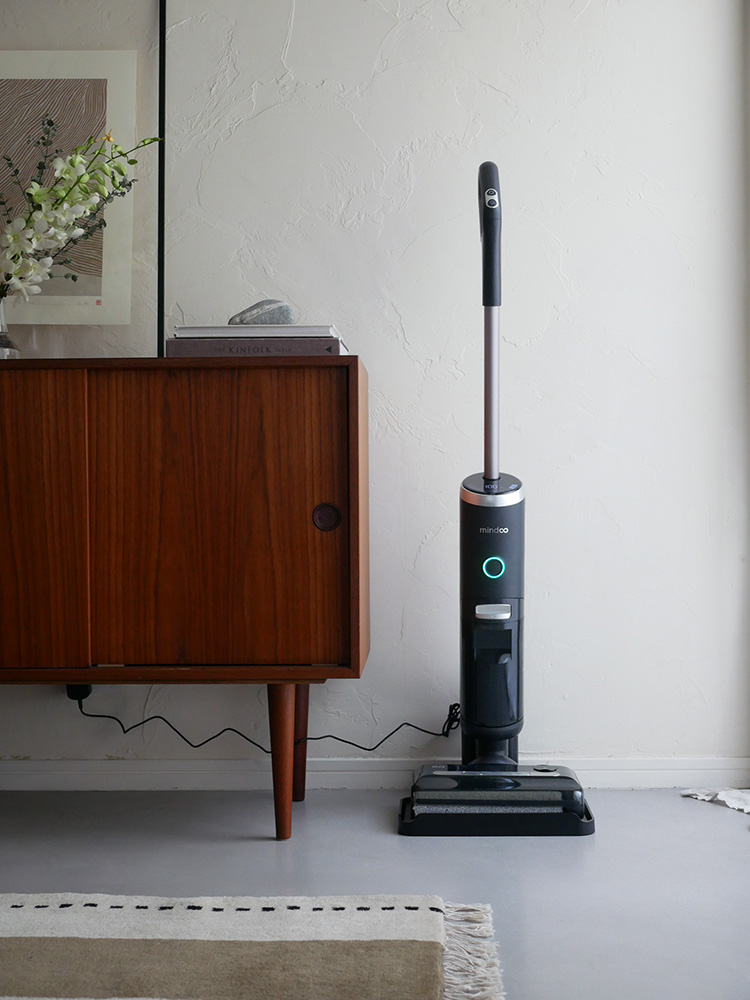 Mindoo AquaX掃除機と充電スタンド、ロールブラシ、フィルターなどの付属品が木製フローリングの上に配置されています。
