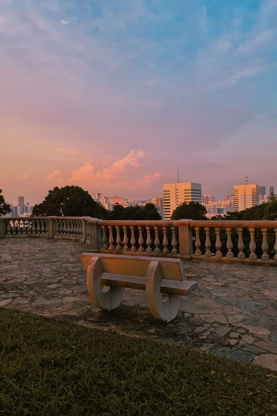 An empty stone bench overlooking a city skyline under a pastel sunset