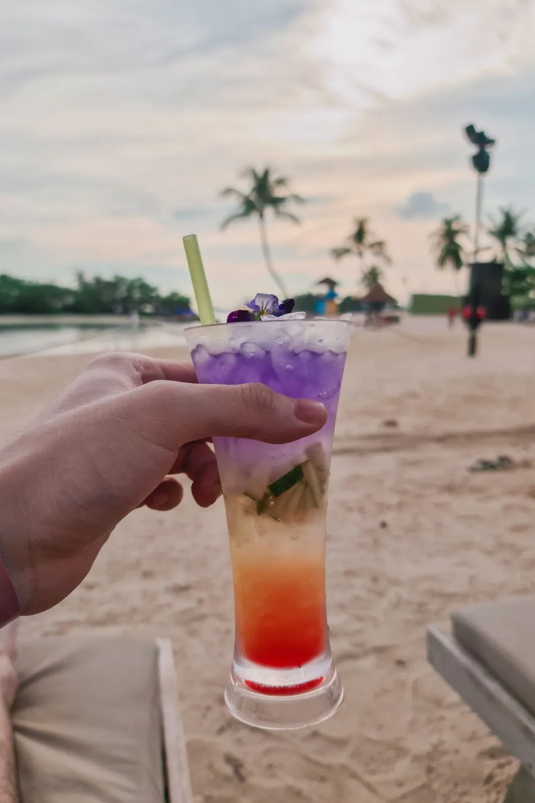 A hand holding a layered purple and red cocktail on a beach