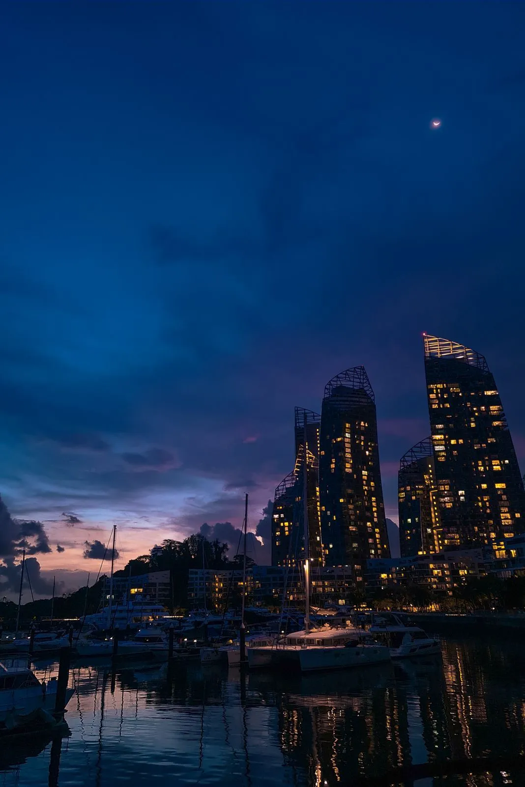 Tall lit skyscrapers rising over a marina with sailboats and a crescent moon