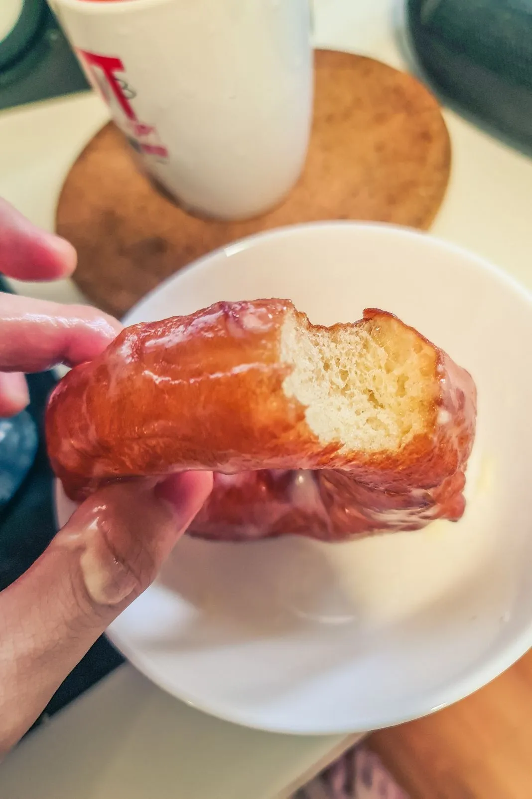 A half-eaten glazed donut held over a plate near a coffee cup