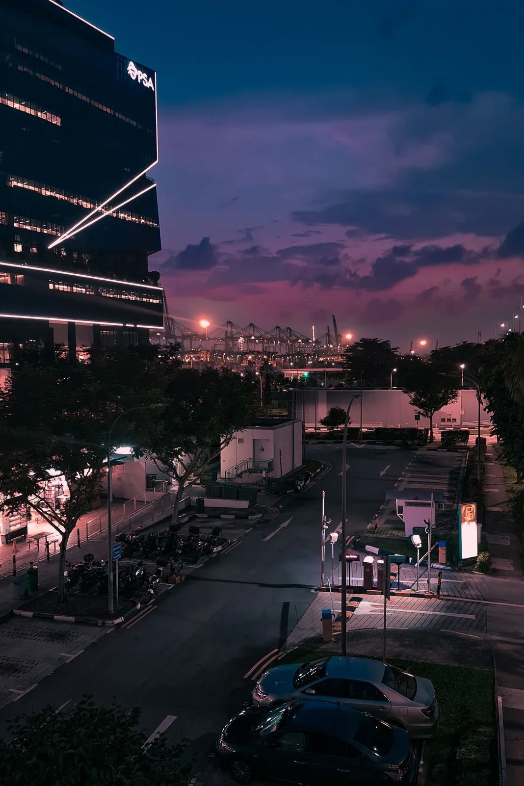 A lit PSA office building beside a parking lot with port cranes in the distance at dusk